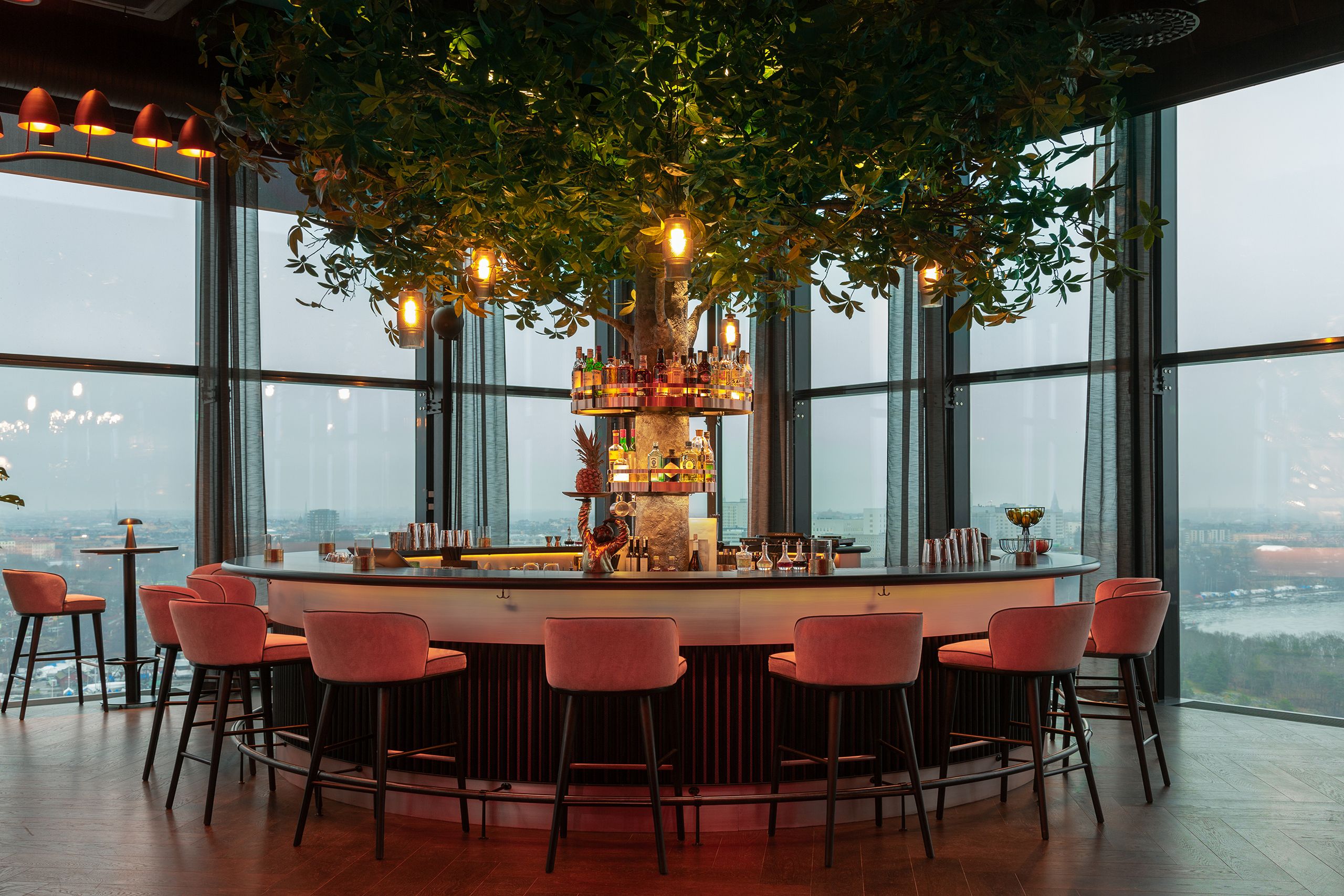 Inmitten des Bildes befindet sich eine atmosphärisch beleuchtete Bar vor einer Panorama Fensterfront.