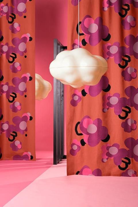 Dekorativer Vorhangstoff von Création Baumann mit grafischem Blumenmuster in Pink- und Violetttönen auf orangem Grund, inszeniert in modernem Interior mit Design-Wolke.