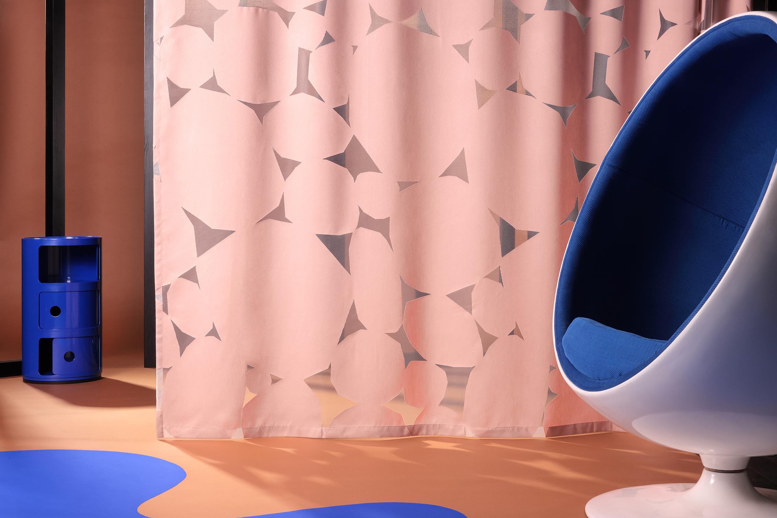 Halbtransparenter Vorhangstoff von Création Baumann in warmem Rosé mit grafischem Muster, präsentiert in zeitgenössischem Wohnraum mit ikonischem Designstuhl und farbigen Accessoires.